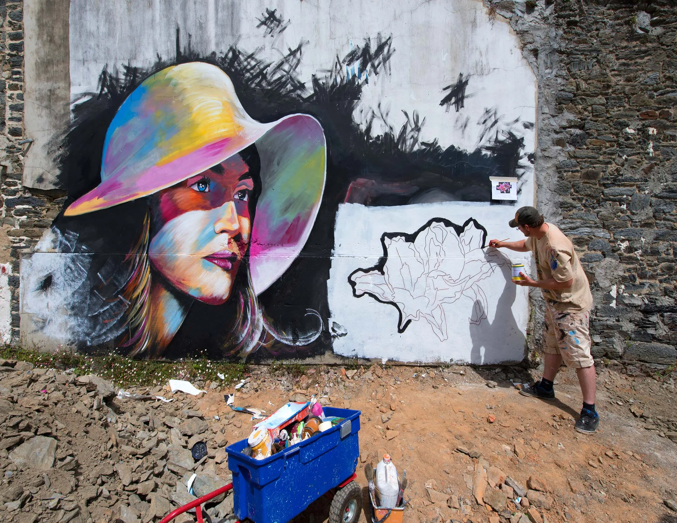Artiste graffeur peignant une carte de la Bretagne à côté d'un portrait de femme au chapeau coloré sur un mur de pierre à Morlaix, culture street art et fresque murale