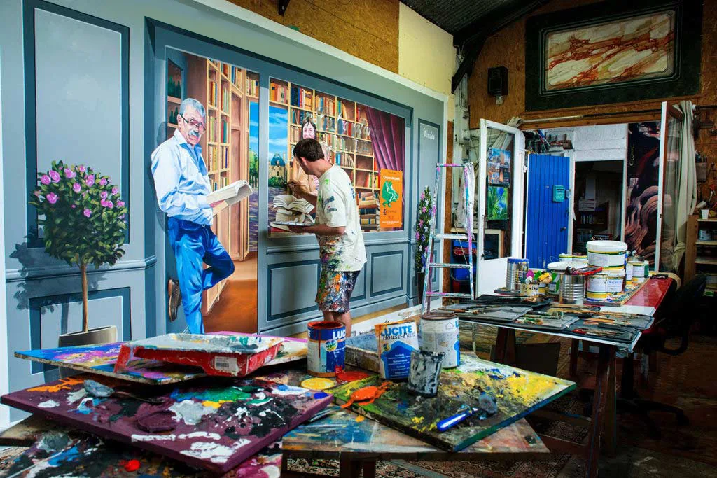 Atelier de Morlaix, peintre devant un trompe-l'œil de bibliothèque, pots de peinture au premier plan