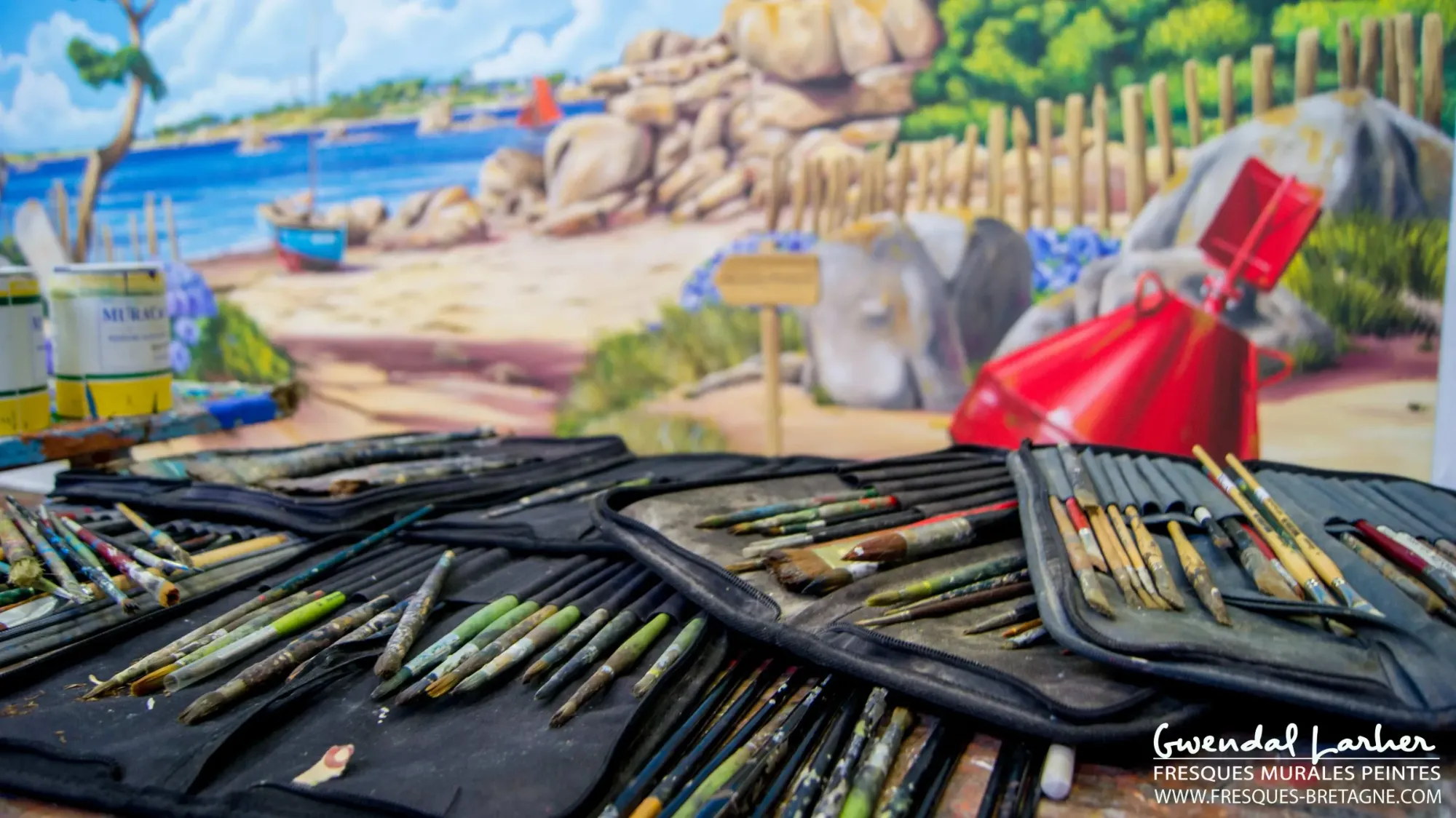 Pinceaux et trousses sur la table d'atelier, devant une fresque de plage et bouée rouge