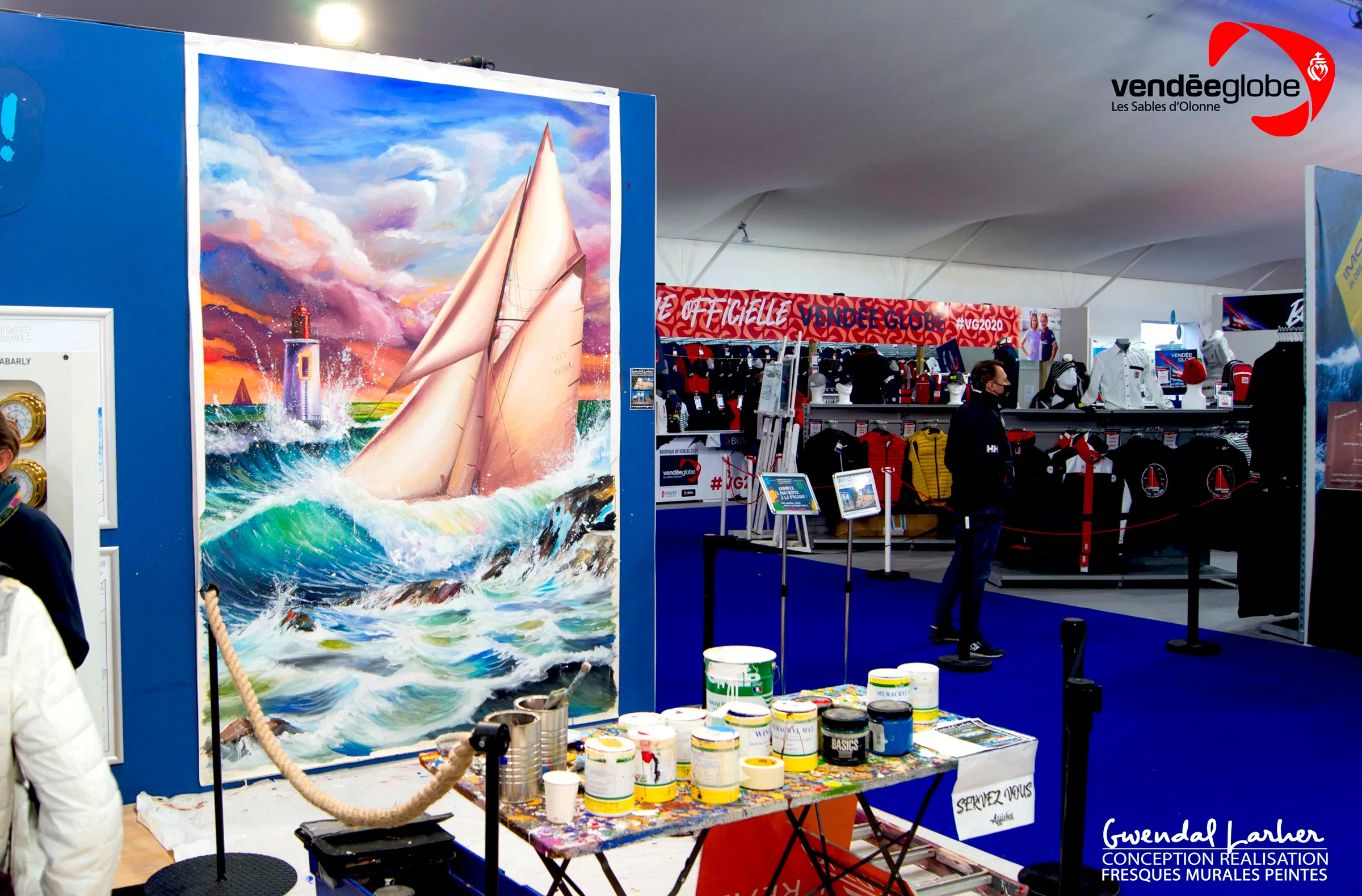 Stand du Village Vendée Globe habillé d'une grande toile peinte d'un voilier sur les vagues, avec les pots de peinture de l'artiste au pied du stand